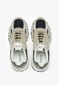 Beżowe i czarne sneakersy z siateczkowymi wstawkami, sznurówkami z przodu i gumowymi podeszwami. Zawierają zielone akcenty na pięcie oraz wzorzasty uchwyt z tyłu.