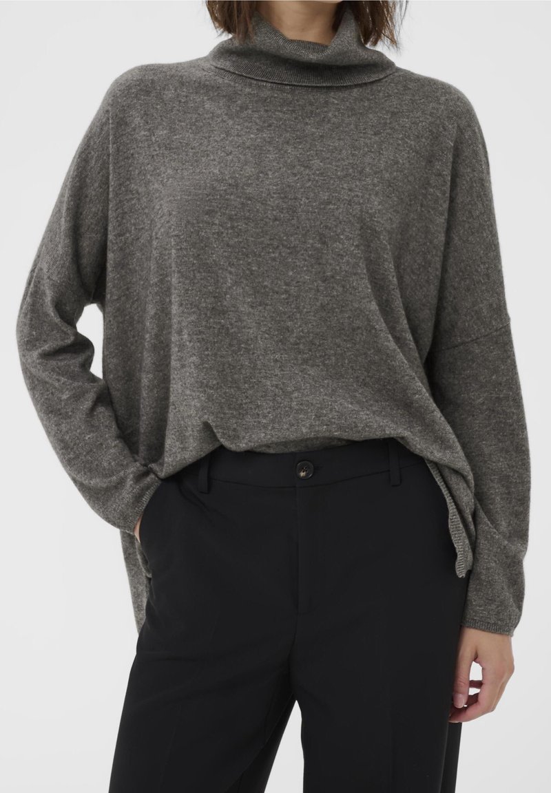 Femme portant un pull col roulé gris ample, rentré dans un pantalon noir taille haute, avec une main dans la poche.