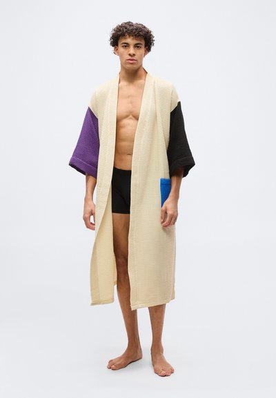 Νεαρός άντρας που στέκεται ξυπόλητος, φορώντας μια μακριά μπεζ ρόμπα με μωβ και μαύρα μανίκια και μια μπλε τσέπη, και από κάτω μαύρα σορτς.