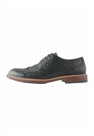 Schwarzer Herren-Brogue-Schuh aus Leder mit Wingtip-Design, Perforationen, Schnürverschluss und gestapeltem Holzabsatz vor weißem Hintergrund.