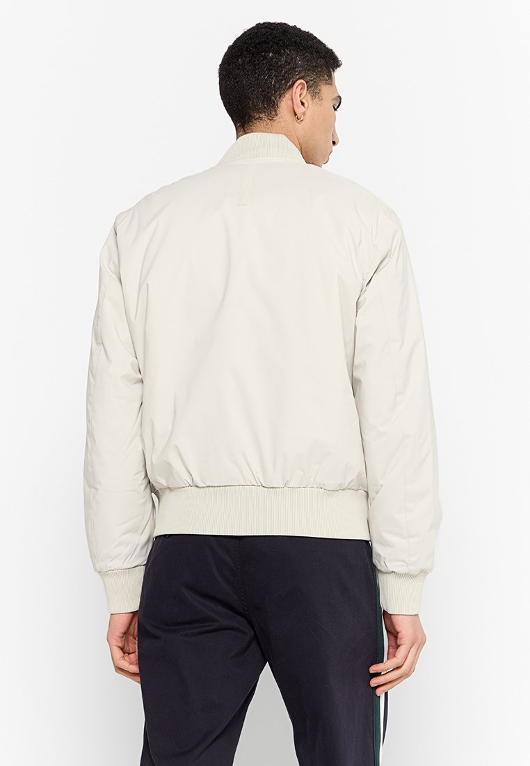 Giacca bomber beige chiaro con colletto e polsini a coste, caratterizzata da una texture liscia e leggermente lucida e da un orlo elasticizzato.