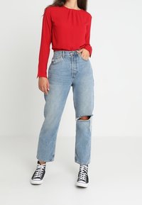 Blusa rossa a maniche lunghe con una vestibilità rilassata, abbinata a jeans a vita alta di colore blu chiaro con ginocchio strappato. Sneakers nere alte.