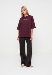 Maroon oversized t-shirt with short sleeves, featuring white embroidered text "LeGer Studio." Paired with black wide-leg trousers and light shoes.