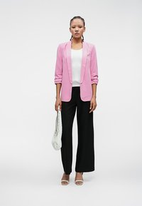 Ružový blazer, biele tričko, čierne široké nohavice a svetlé sandále. Modelka drží bielu tašku. Hladká tkanina, strih na mieru, ležérny vzhľad.