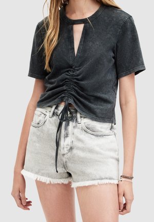 Kobieta ubrana w czarny, sprany, marszczony krótki t-shirt z dekoltem w kształcie łezki oraz jasnoszare, postrzępione krótkie spodenki dżinsowe, z bransoletkami na nadgarstku.