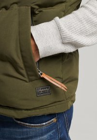 Olivgrüne gepolsterte Weste mit einer Reißverschlusstasche, einer Leder-Ziehschlaufe und einem gewebten grauen Pullovern. Verfügt über ein Markenetikett in der Nähe des Saums.