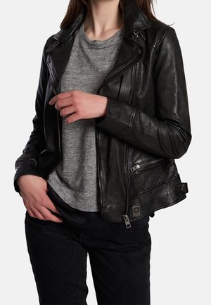 Mujer con chaqueta de cuero negra sobre camiseta gris y pantalones negros, manos ajustando la cremallera de la chaqueta y apoyadas en la cadera.