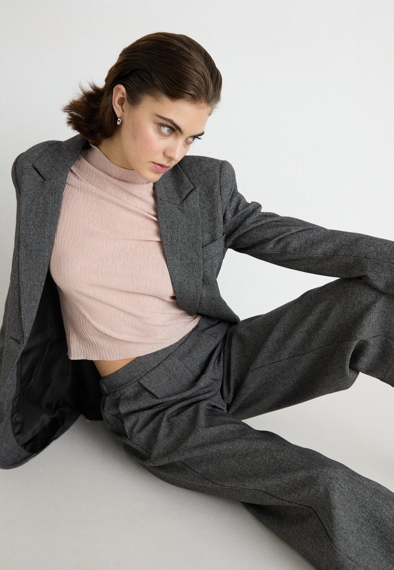 Chaqueta de traje gris ajustada y pantalones de pierna amplia combinados con un crop top de canalé rosa claro. Textura de tela suave, diseño minimalista.