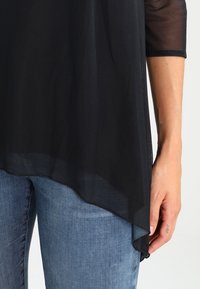 Schwarzes transparentes Oberteil mit lockerer Passform, asymmetrischem Saum und kurzen Netzärmeln, kombiniert mit hellblauen Denim-Jeans.