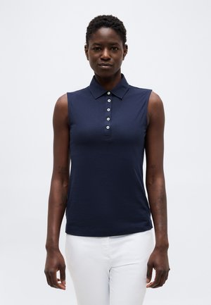 Lühikeste juustega naine, kes kannab varrukateta tumelõherohelist polo-särki ja valgeid pükse, seisab ühevärvilise heleda tausta ees.