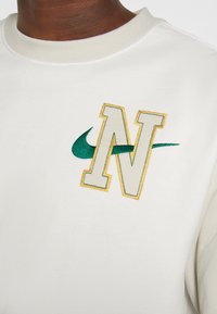 Vit sweatshirt med en broderad "N"-logotyp i beige och gult, samt en grön Nike-swoosh. Texturerad tyg med rund halsringning.