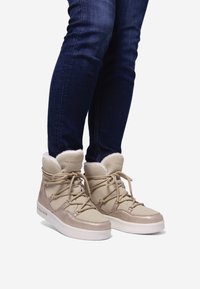 Botas de invierno beige con una parte superior texturizada, forro de piel sintética y suela de goma blanca robusta. Diseño con cordones y detalle del logo en el lateral.