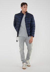 Giovane uomo in piedi, indossa un giubbotto blu scuro, una maglietta grigia, dei jeans grigio chiaro e delle sneakers bianche con dettagli blu.