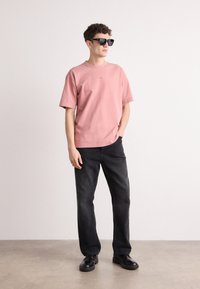 Lyserød bomulds-t-shirt, afslappet pasform, parret med mørkegrå denimbukser. Sorte firkantede solbriller og sorte sko fuldender looket.