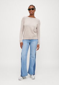Pull en tricot beige avec un col rond et un bas côtelé, associé à un jean bootcut bleu clair avec des accents sur les panneaux latéraux, et des baskets grises.