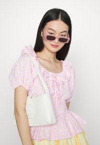 Blusa riscada a rosa com padrões florais amarelos, gola com babados e mangas bufantes. Bolsa branca com formato retangular e detalhe de fivela.