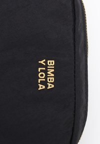 Bolsa de tela negra con el texto dorado en relieve "BIMBA Y LOLA" en la parte frontal. La textura es suave con un leve brillo. Cierre con cremallera a lo largo del borde.