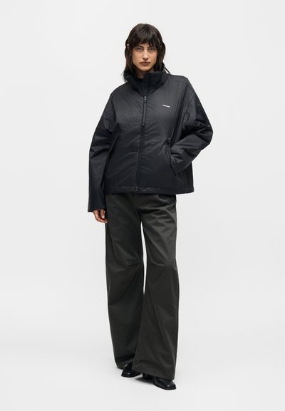 Γυναίκα όρθια με το χέρι στην τσέπη, φοράει μαύρο μπουφάν με φερμουάρ, φαρδιά σκούρα παντελόνια και μαύρα παπούτσια σε απλό φόντο.