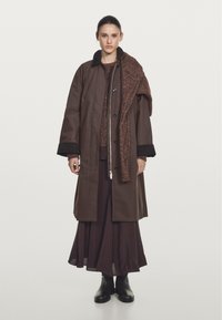 Brown long coat with a black collar, worn over a maroon textured sweater and a brown skirt. Black ankle boots complete the outfit.