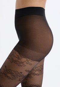 Zwarte panty heeft een gladde, transparante textuur met een bloemenkantpatroon aan de onderkant en een klein stippenontwerp op het bovenste gedeelte.