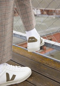 White Veja sneakers with green accents and logo, textured sole, paired with patterned brown trousers and white socks, positioned on wooden surface.