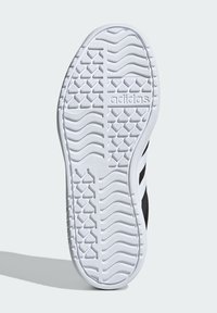 Semelle de chaussure de sport noire avec une semelle en caoutchouc blanc texturé, présentant un motif en vagues pour l'adhérence et un détail de logo en relief.