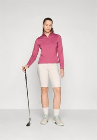 Lichtroze golfshirt met lange mouwen en een halve ritssluiting, gecombineerd met beige shorts en lichtgekleurde golfschoenen. Houdt een golfclub vast.