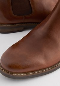 Bottes cheville en cuir marron avec une finition lisse, des panneaux latéraux élastiques et une semelle robuste avec un détail de couture décorative.