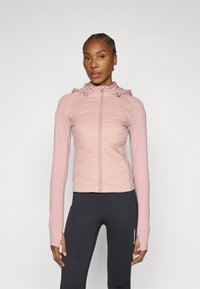 Világos rózsaszín cipzáras kabát kapucnival, a mellkas területén steppelt mintával, hosszú ujjakkal és hüvelykujjnak kialakított nyílásokkal. Fekete leggingssel párosítva.