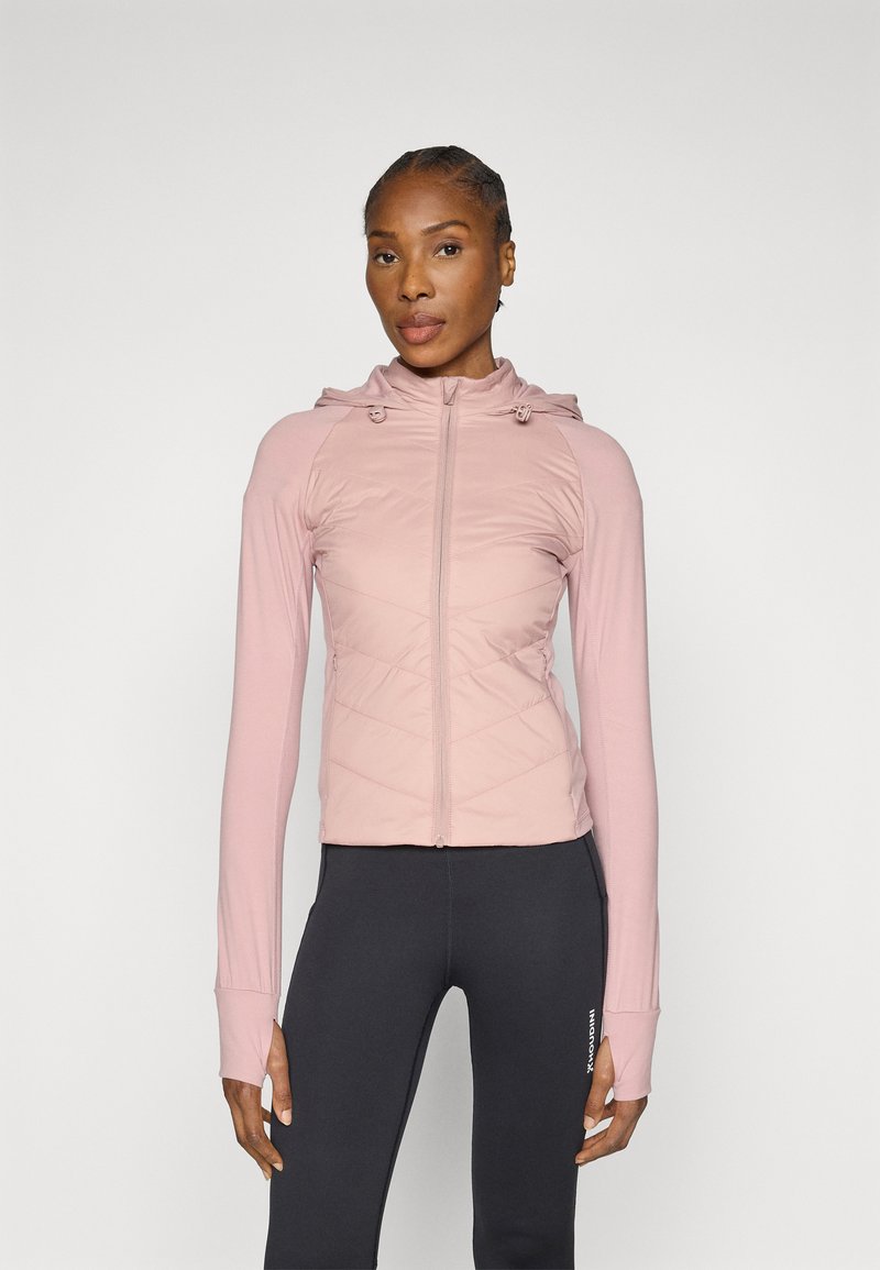 Világos rózsaszín cipzáras kabát kapucnival, a mellkas területén steppelt mintával, hosszú ujjakkal és hüvelykujjnak kialakított nyílásokkal. Fekete leggingssel párosítva.