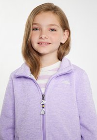 Chaqueta de forro polar de color púrpura claro con un acabado texturizado, que presenta un cuello alto y un tirador de cremallera decorativo. Camiseta de rayas debajo.