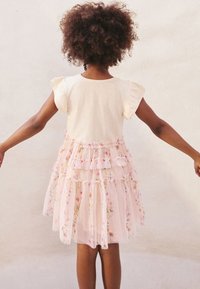 Mouwloze jurk met een lichtbeige top, voorzien van een gelaagde roze tule rok versierd met bloemmotieven en gerimpelde details.