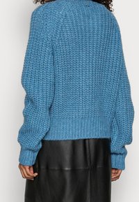 Oversized modrý pletený svetr s ribbovým vzorem, sestupnými rameny a rovním lemem. Obsahuje malý stříbrný detail na zadní straně.