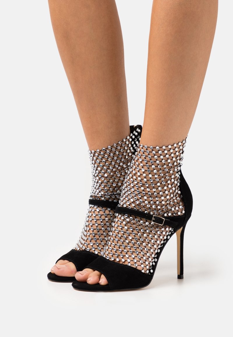 Sandales à talons hauts noires portées avec des chaussettes résille à la cheville ornées de clous argentés sur les pieds, sur fond blanc.
