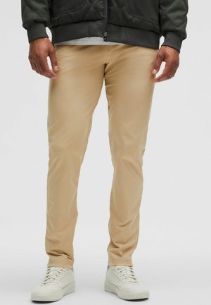 Beżowe spodnie o dopasowanym kroju wykonane z gładkiego materiału, z wąskim fasonem, zestawione z białymi trampkami. Prosty i funkcjonalny design.
