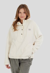Kvinde iført en cremet farvet fleece-pullover med trykknapper og grønne kordfløjlsbukser, stående med den ene hånd i lommen.
