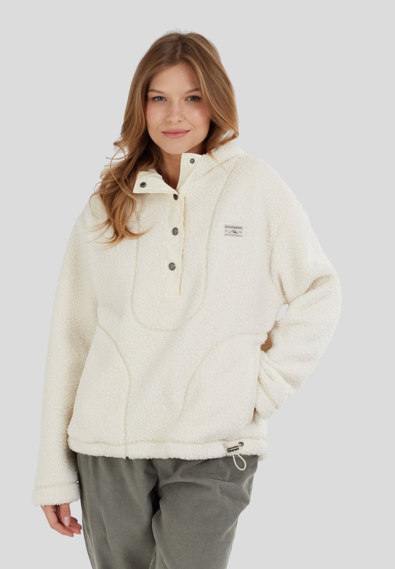 Kvinde iført en cremet farvet fleece-pullover med trykknapper og grønne kordfløjlsbukser, stående med den ene hånd i lommen.