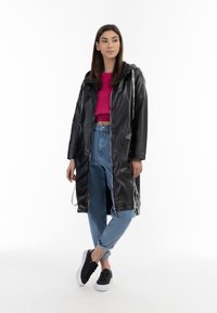 Zwarte waterdichte regenjas met een ritssluiting aan de voorkant, een capuchon en aantrekkoorden, gedragen over een cropped roze top en lichtblauwe jeans, gecombineerd met zwarte sneakers.