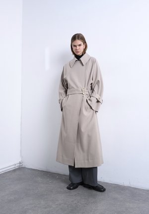 Femeie stând în picioare lângă un perete alb, purtând un trench bej cu centură, un pulover negru cu guler înalt, pantaloni largi gri și pantofi negri.