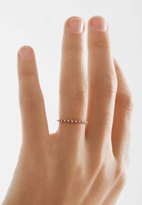 Anillo de banda delgada de oro rosa, engastado con gemas alternas transparentes y rojas, llevado en una mano con un acabado natural y uñas cortas.