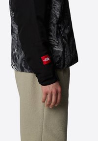 Svart och grå mönstrad jacka med en slät yta, med en röd logotyp på ärmen, bärs över beiga fleecebyxor.