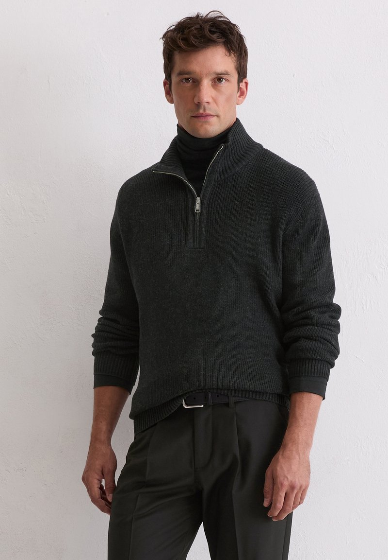 Sweater zippé gris foncé avec un col haut, texture côtelée et manches ajustées, associé à un pantalon noir et une ceinture noire.