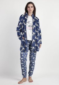 Conjunto de pijama de felpa azul con patrones de Snoopy y Woodstock; incluye pantalones largos y una chaqueta con cuello y bolsillos. Camiseta gráfica blanca debajo.