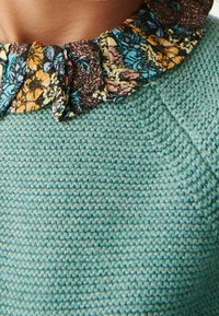 Pletený tykvový svetr s volánkovým květinovým límcem, na kterém jsou vzory v oranžové, modré a černé barvě, zdůrazňující kontrastní textury a vzory.