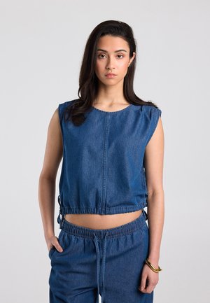 Vrouw met lang donker haar draagt een mouwloze blauwe denim top en bijpassende blauwe denim broek, staand tegen een effen lichte achtergrond.