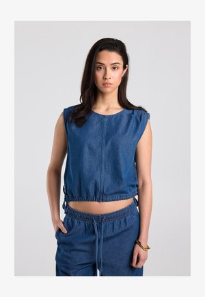 Frau mit langen dunklen Haaren trägt ein ärmelloses blaues Denim-Top und passende blaue Denim-Hosen und steht vor einem einfarbigen hellen Hintergrund.