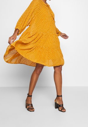 Femme portant une robe fluide jaune à pois et des sandales noires à talons, debout devant un fond blanc uni.
