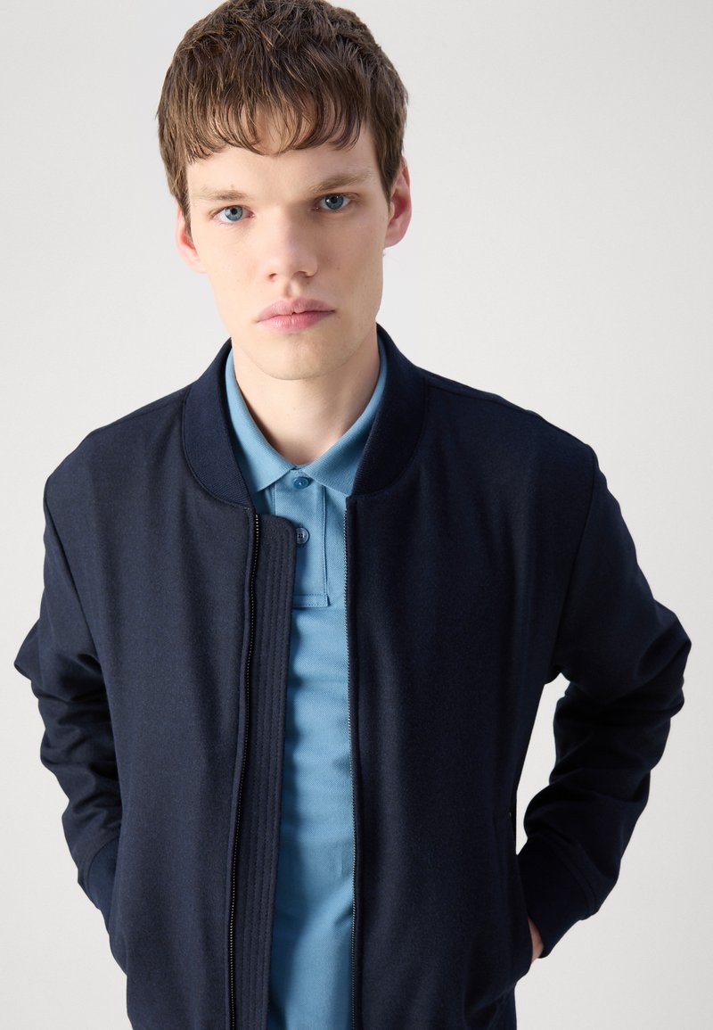 Granatowa kurtka bombowca z zapięciem na zamek, żeberkowym kołnierzem i mankietami, nałożona na jasnoniebieską koszulkę polo z guzikami.