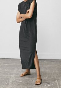 En person står med armarna korsade, iklädd en ärmlös svart midiklänning och bruna sandaler med remmar på en betonggolv.