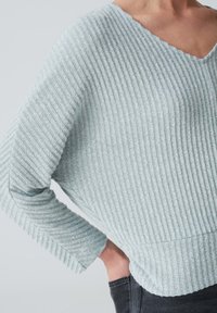 Hellblaues, geripptes Sweatshirt mit V-Ausschnitt und langen Ärmeln, aus strukturiertem Stoff und mit leicht lockerer Passform. Schwarze Hosen sind sichtbar.